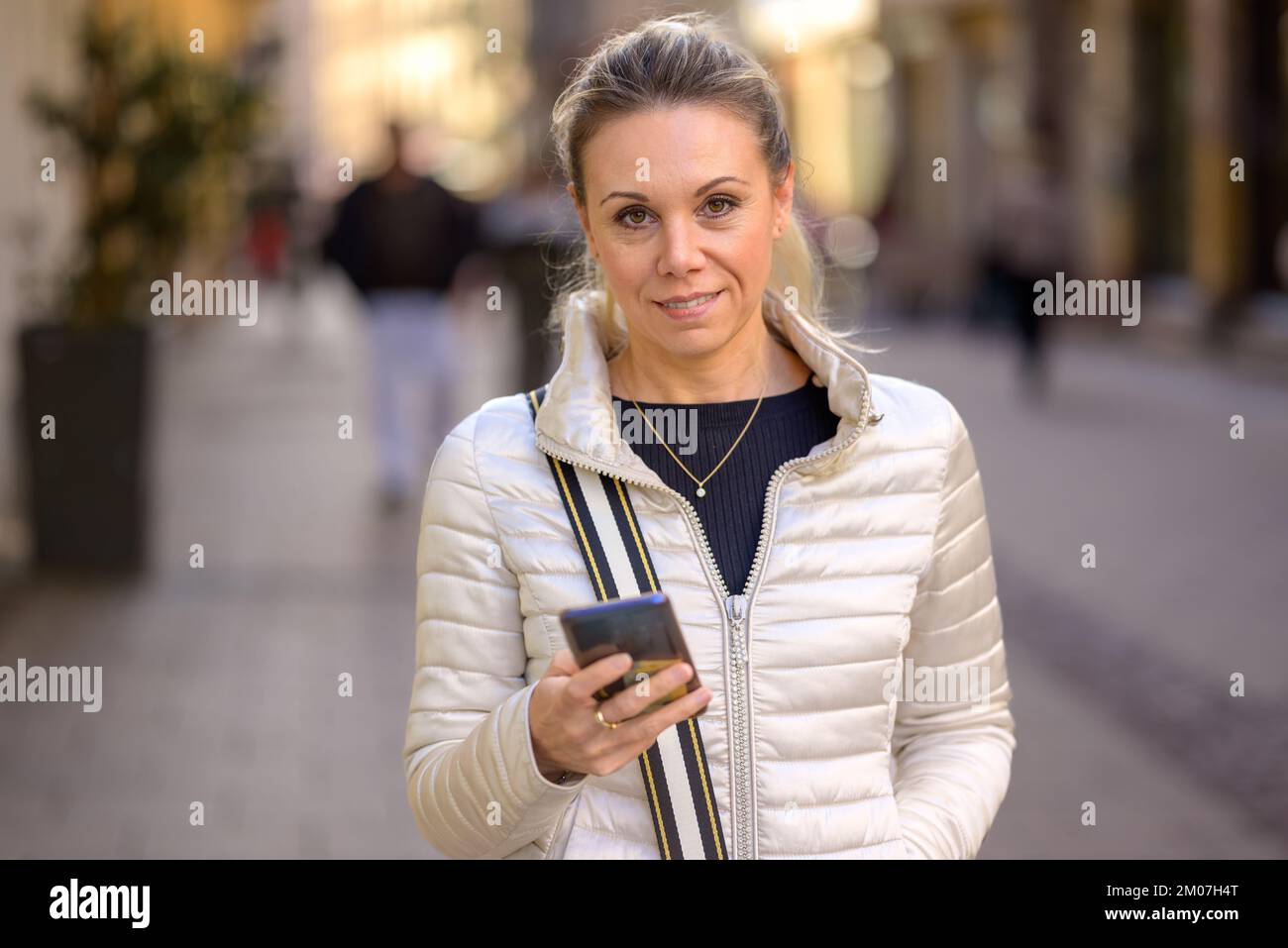 Upper body portrait of a smiling middle aged woman with blonde hair and a ponytail looking at the camera while typing on her cell phone in a shopping Stock Photo