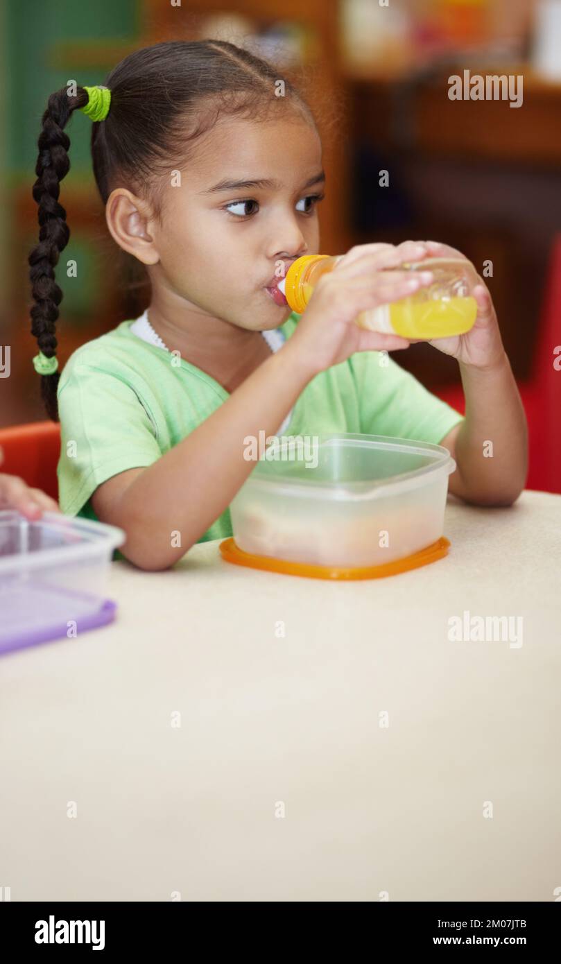 Model behaviour. Preschool ethnic girl sitting at a table with her sandwich and drinking her juice. Stock Photo