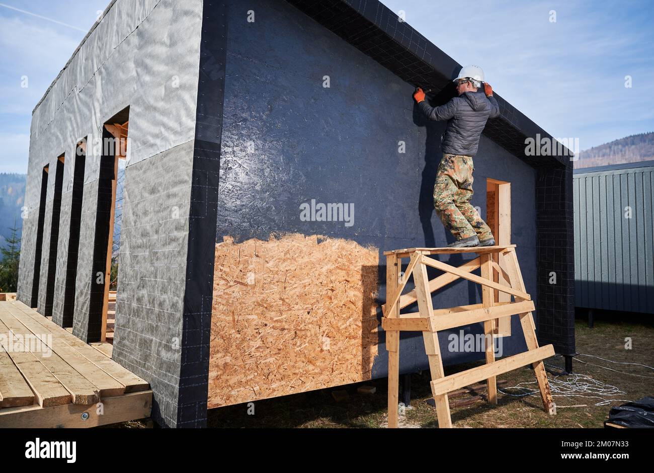 Male painter using paint roller, doing exterior paint work in a black color. Man worker building wooden frame house. Carpentry and construction concept. Stock Photo