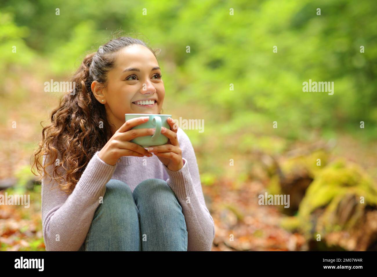 Happy woman sitting in a forest drinking coffee looking away Stock Photo