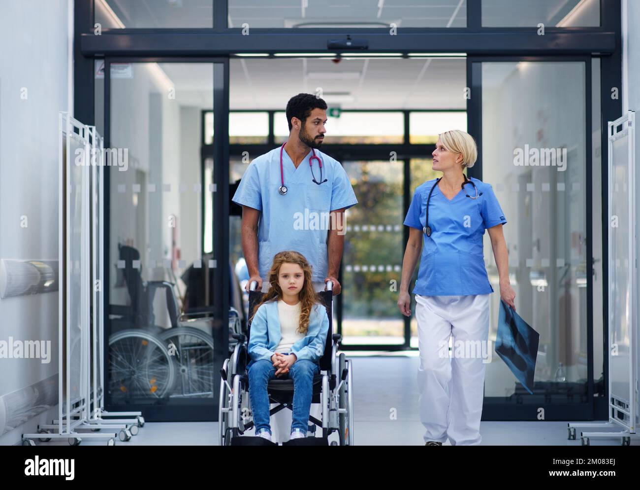 Pregnant doctor with her colleague pushing litle girl at wheelchair at pediatric department. Stock Photo