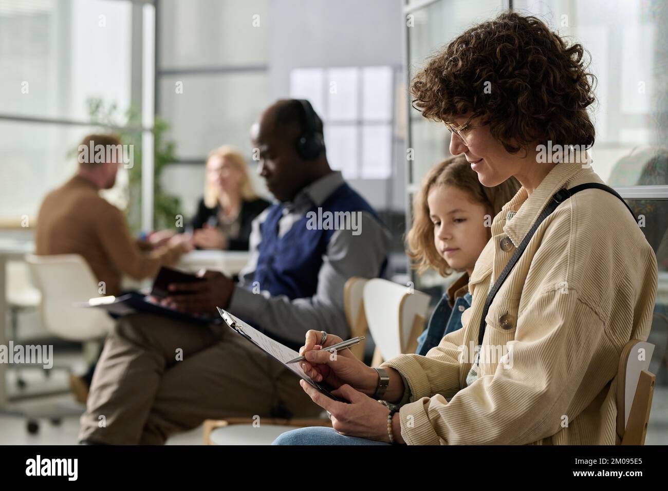 Side view portrait of young mother and daughter filling application forms while waiting in line at immigration office, copy space Stock Photo