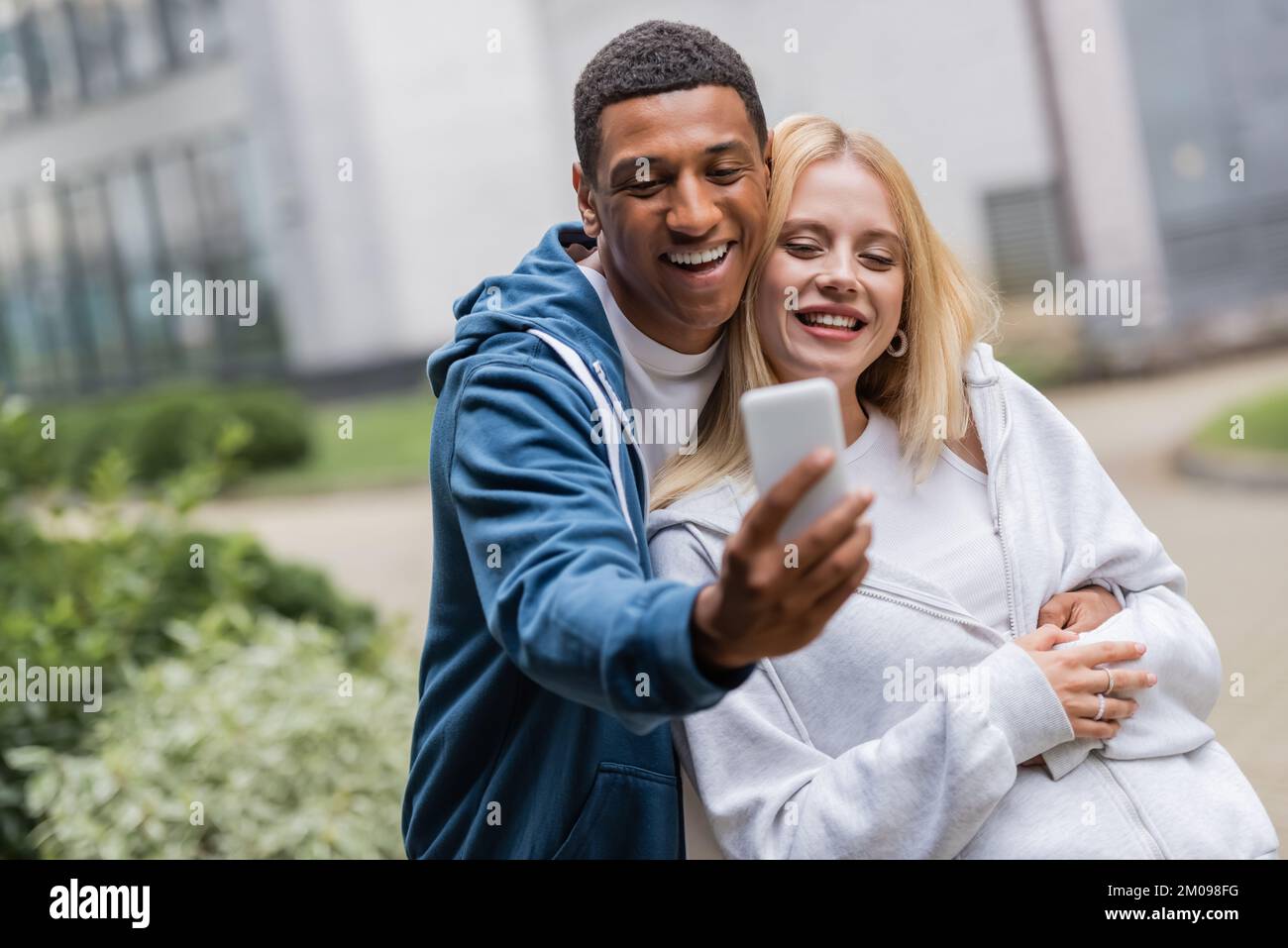 cheerful interracial couple in hoodies taking selfie on mobile phone outdoors Stock Photo