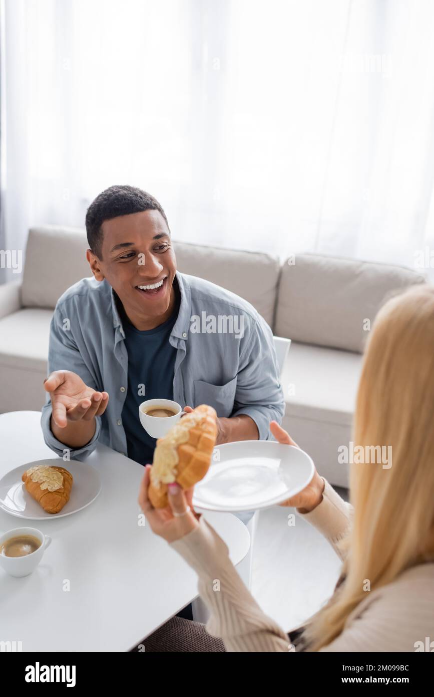 happy african american man with coffee cup talking to blonde girlfriend holding tasty croissant Stock Photo