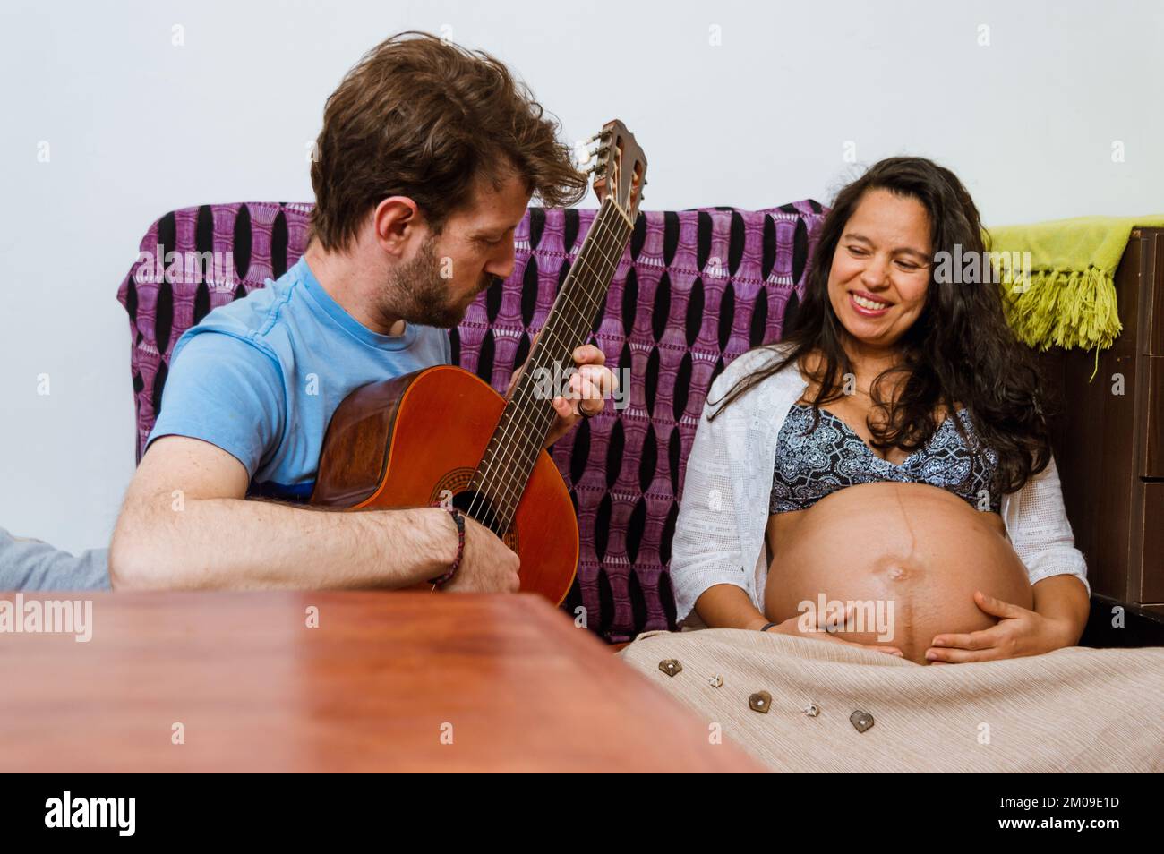 adult caucasian argentinian man playing the acoustic guitar for his baby that is inside the womb of his wife, a brunette brazilian adult woman, sittin Stock Photo
