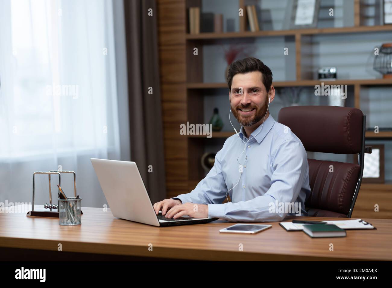 Portrait of successful businessman investor inside office, mature man smiling and looking at camera using laptop at work and wearing white headphones for video call, Stock Photo
