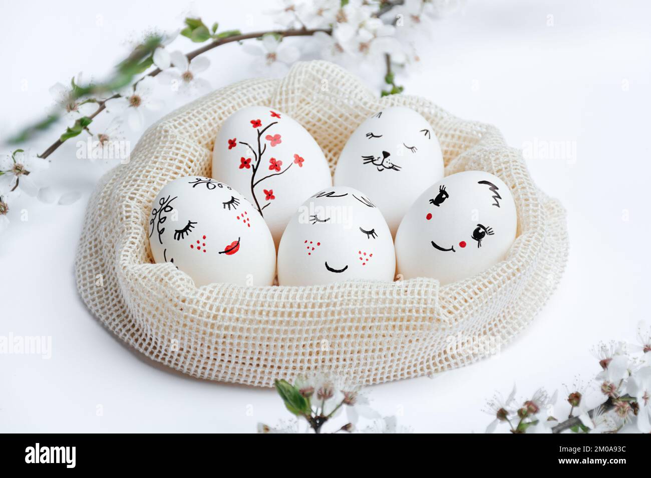 Defocused focus on foreground flowering branch. Easter delicately painted eggs in eco-friendly mesh bag on white table. Funny faces, flowers, viburnum are painted on eggs. Handmade workshop Stock Photo