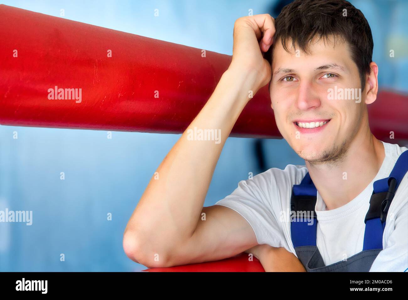 Young Caucasian worker smiles and looks into camera. Portrait of Caucasian man 30 years old. Leaning on red pipe inside room. Genuine happy worker. Stock Photo
