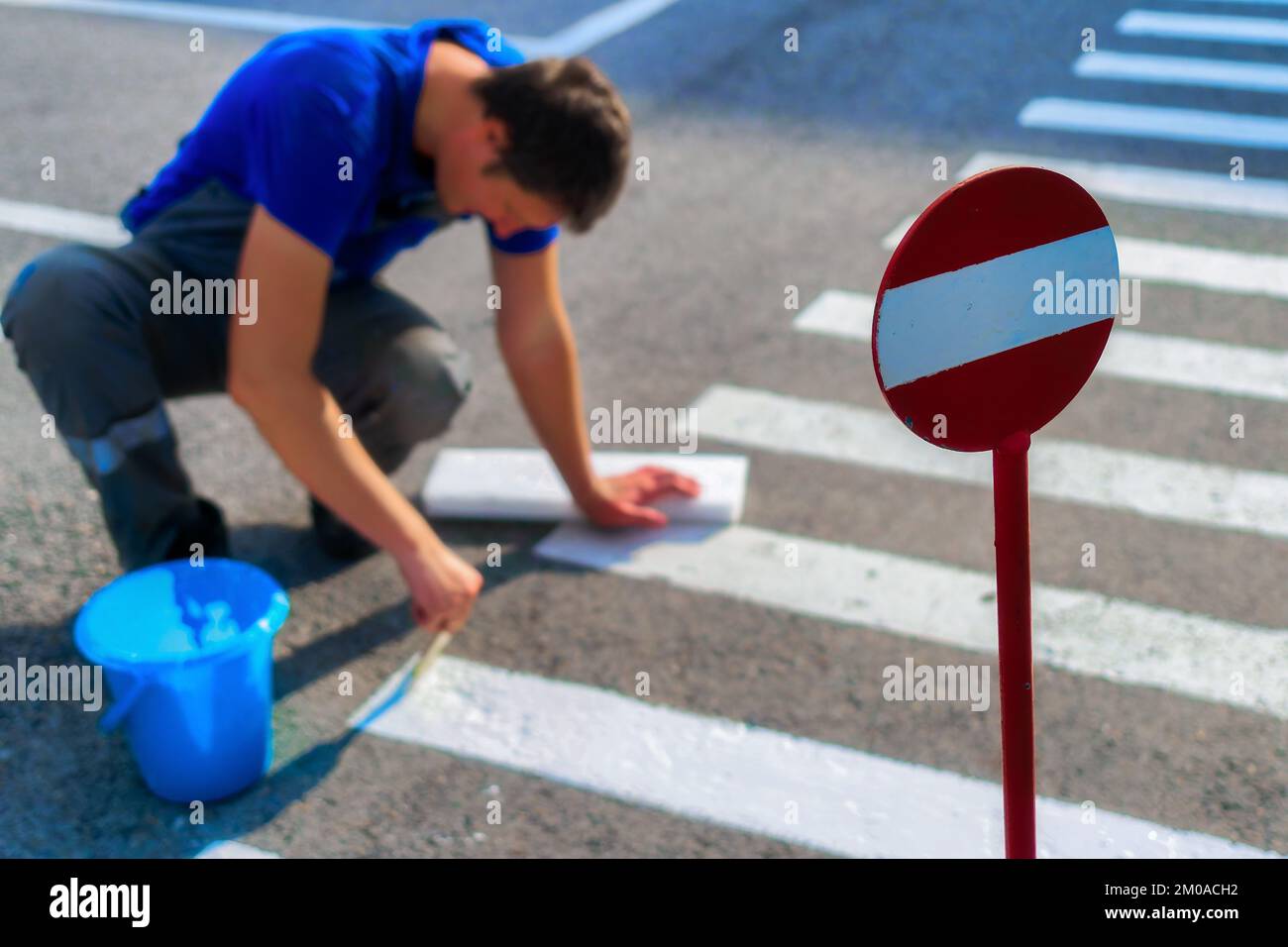 Road service worker sits on his lap and paints striped pedestrian crossing on asphalt with brush. Markings for pedestrians on roadway. Safe transition for people. Real workflow.. Stock Photo