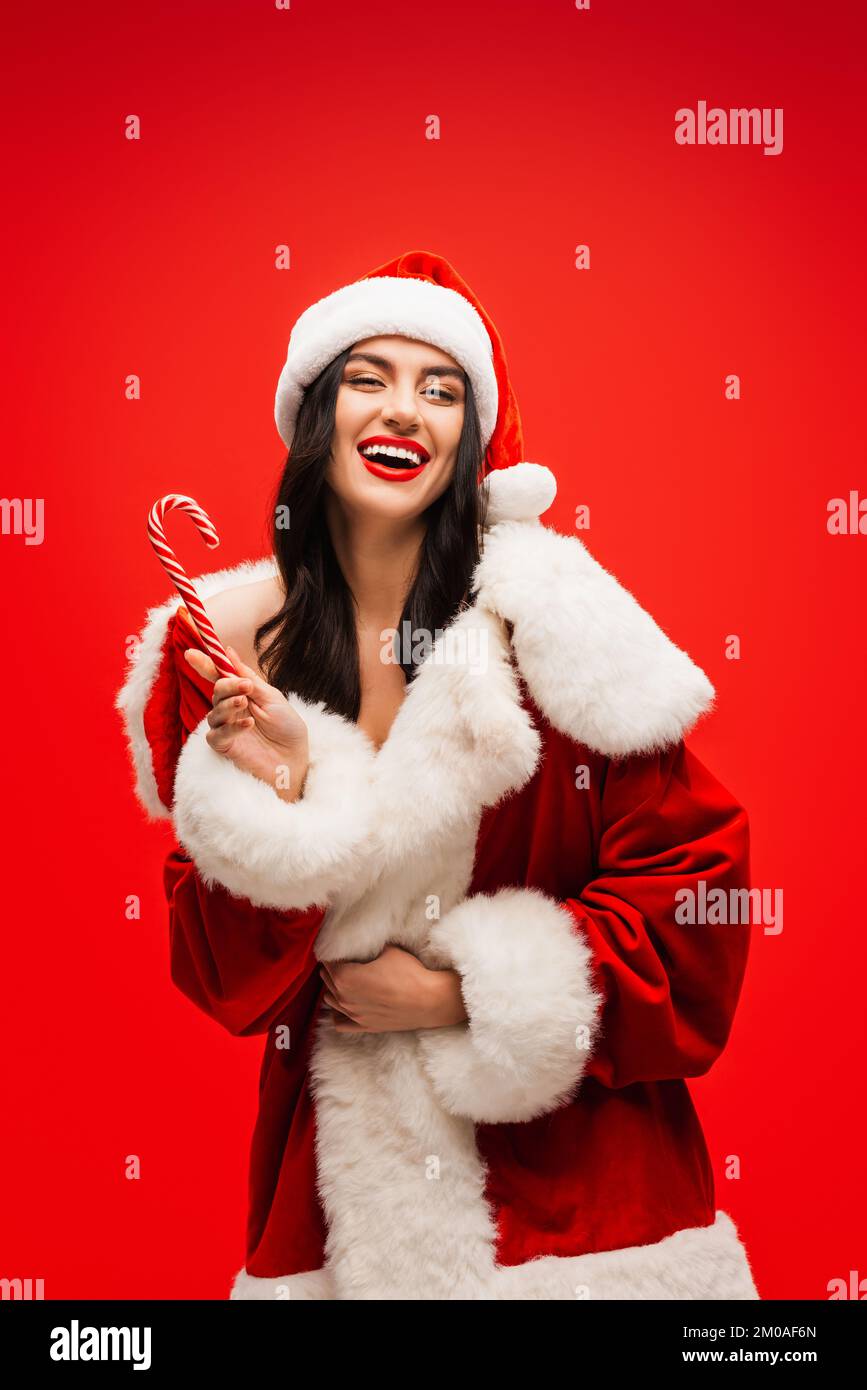 Cheerful young model in santa costume holding striped lollipop isolated on red Stock Photo
