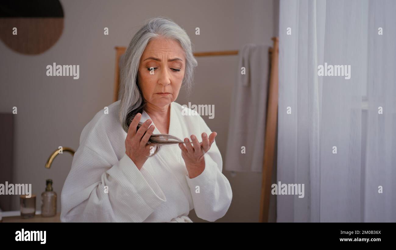 Caucasian old sad senior mature woman lady grandmother model in bathrobe in bath touching gray long smooth thin hair worry about lost hairline weak Stock Photo