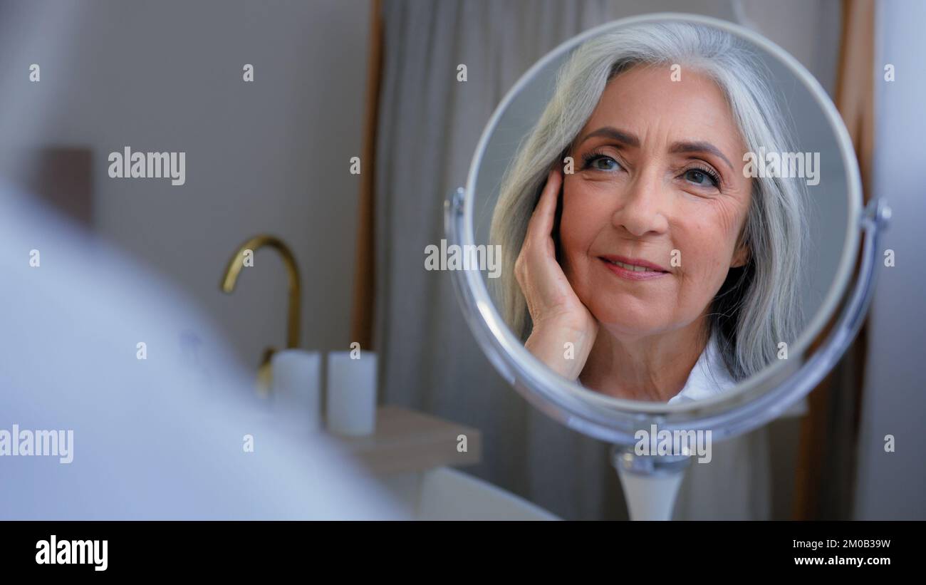 Pensive 50s middle-aged Caucasian woman senior touching face skin looking at mirror reflection smiling enjoy soft facial. Older lady pampering healthy Stock Photo