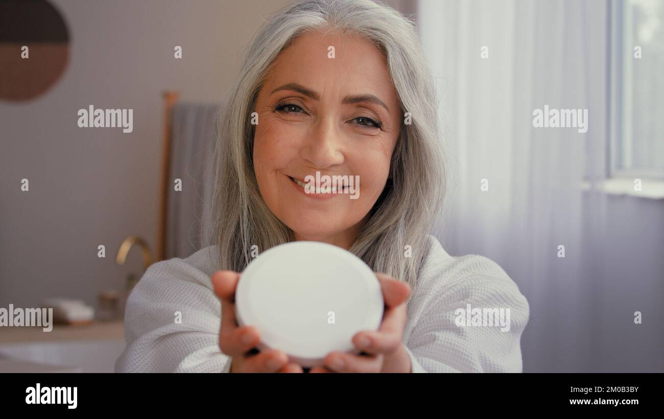Happy old Caucasian 50s senior model gray-haired mature woman smile toothy 60s lady in bathroom showing jar of anti-aging cosmetic natural organic Stock Photo