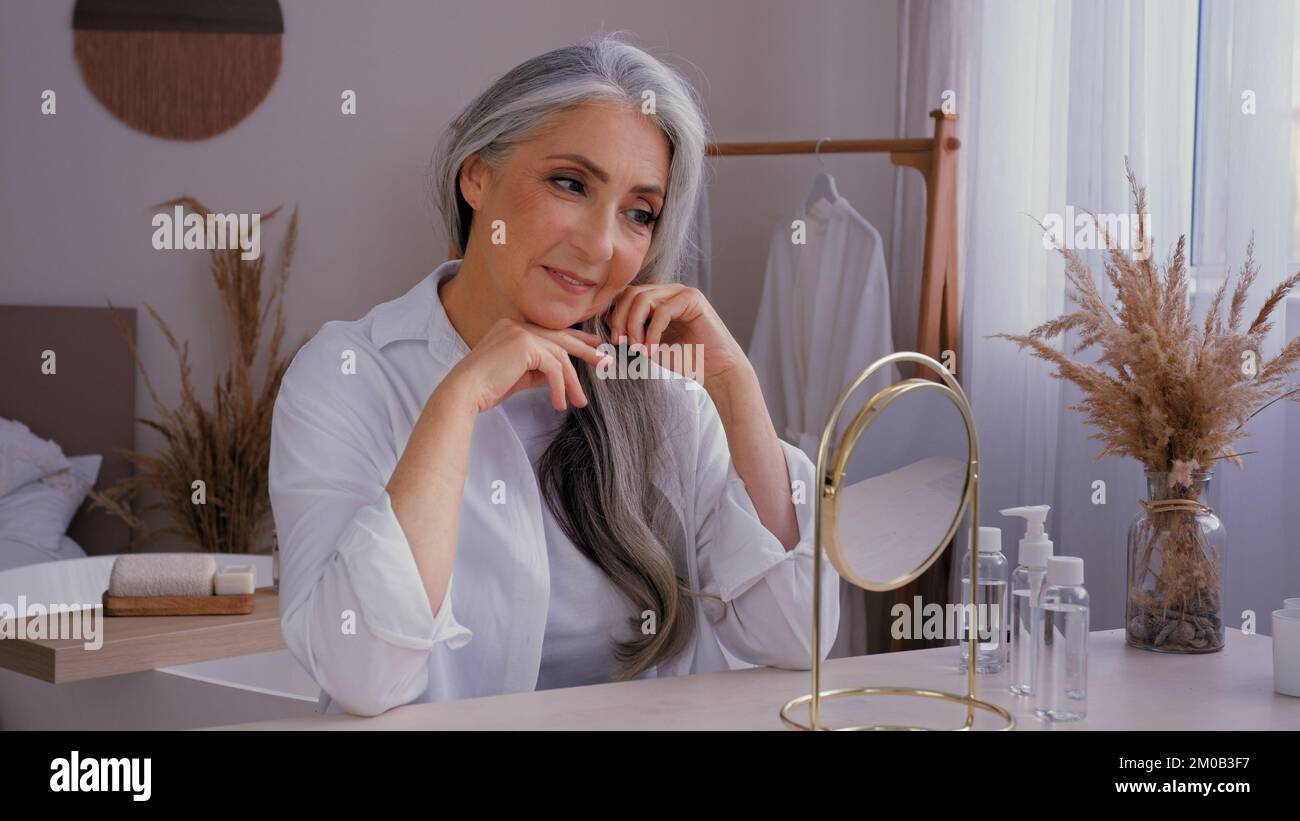 Caucasian 60s aged senior mature lady old gray-haired 50s woman female looking at mirror reflection pampering touching wrinkled face anti-aging Stock Photo