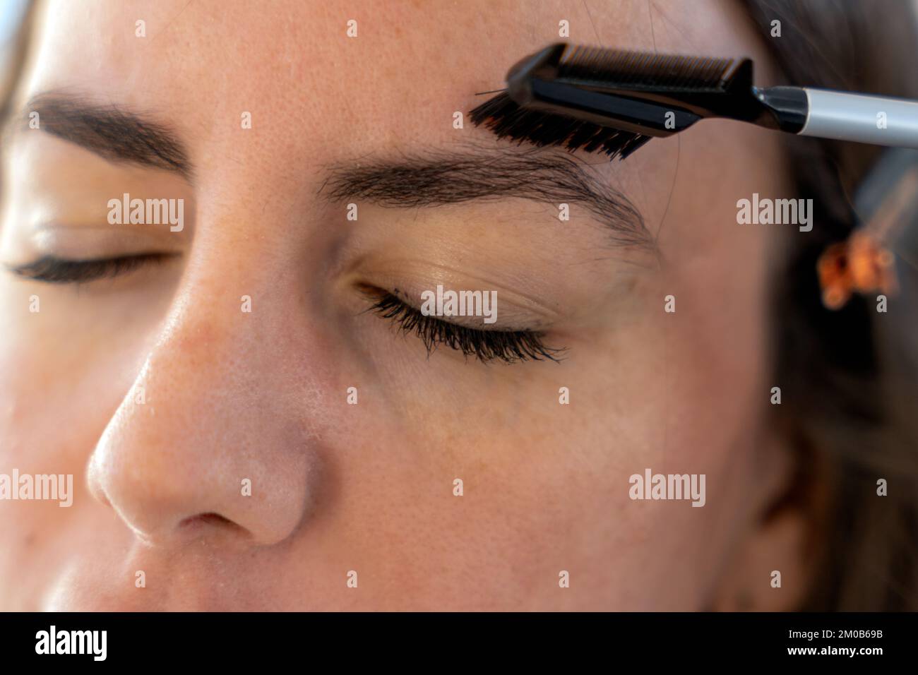 Closeup of crop young female with closed eyes brushing eyebrows with comb while doing makeup in