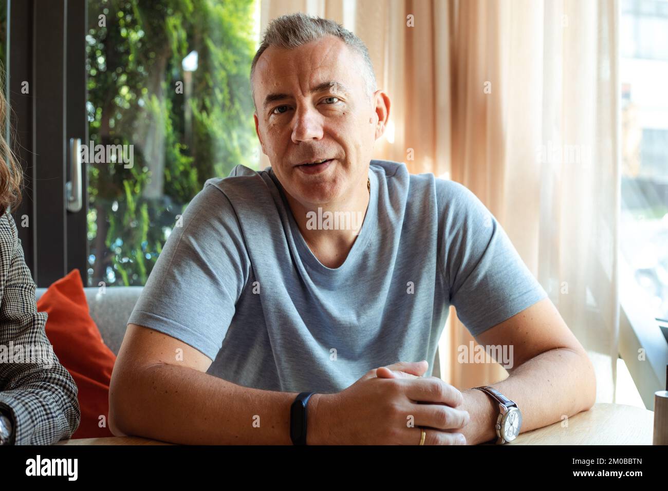 Smiling confident grizzled man in gray t-shirt, with smartwatch, look at camera, sit at table near window. Interview job Stock Photo