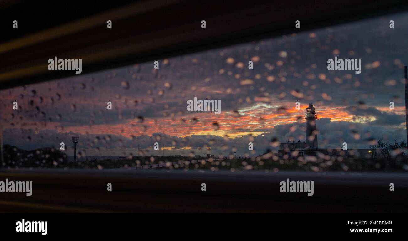 Looking out at sunrise from the inside of a campervan on a rainy day through the blinds towards Flamborough Head lighthouse and beacon on the headland Stock Photo
