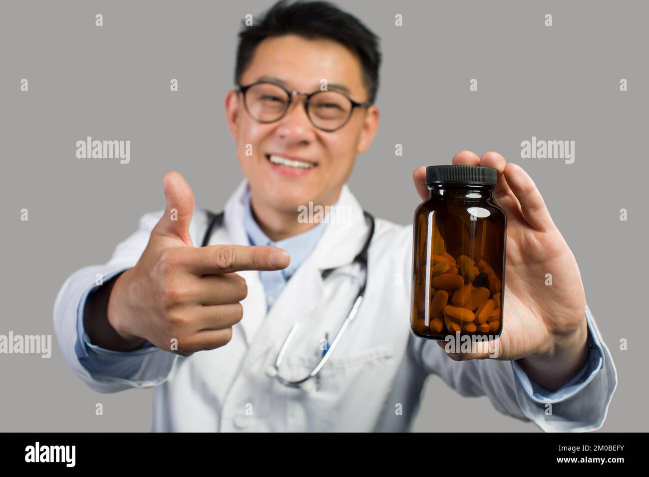 Smiling adult chinese male therapist in white coat and glasses points finger at jar of medicines