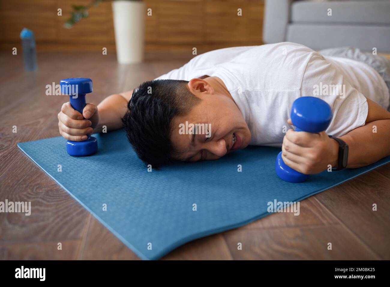 Tired asian mature man lying on fitness mat with dumbbells in hands, resting after domestic workout in living room Stock Photo