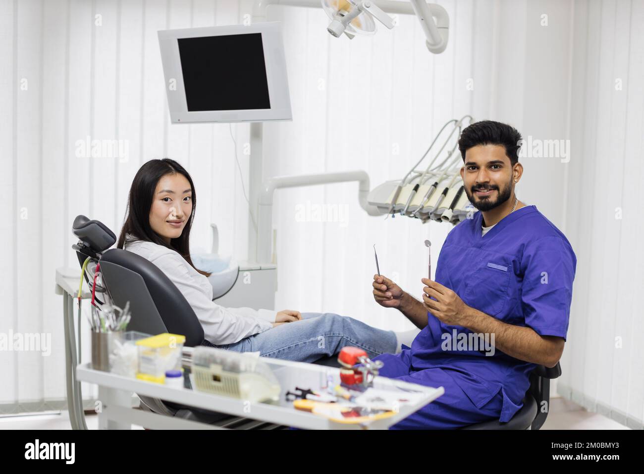 Teeth treatment in dental hospital. Smiling beautiful asian woman sitting in the dentist's chair in light modern dental clinic with young attractive man dentist with tools and looking at camera. Stock Photo