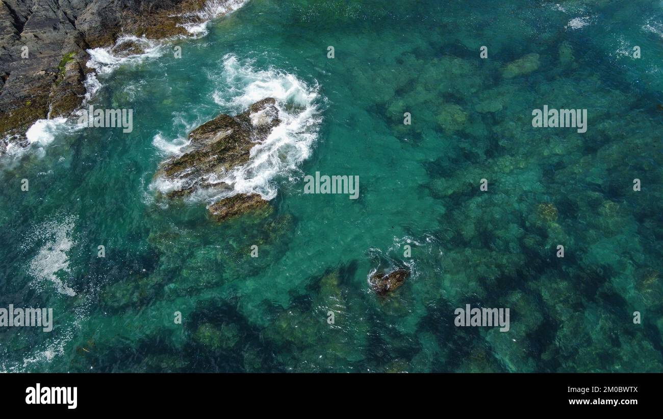 Black moss-covered rocks among turquoise waves of the Celtic Sea. White sea foam on the waves. Waters of the Atlantic Ocean. Stock Photo