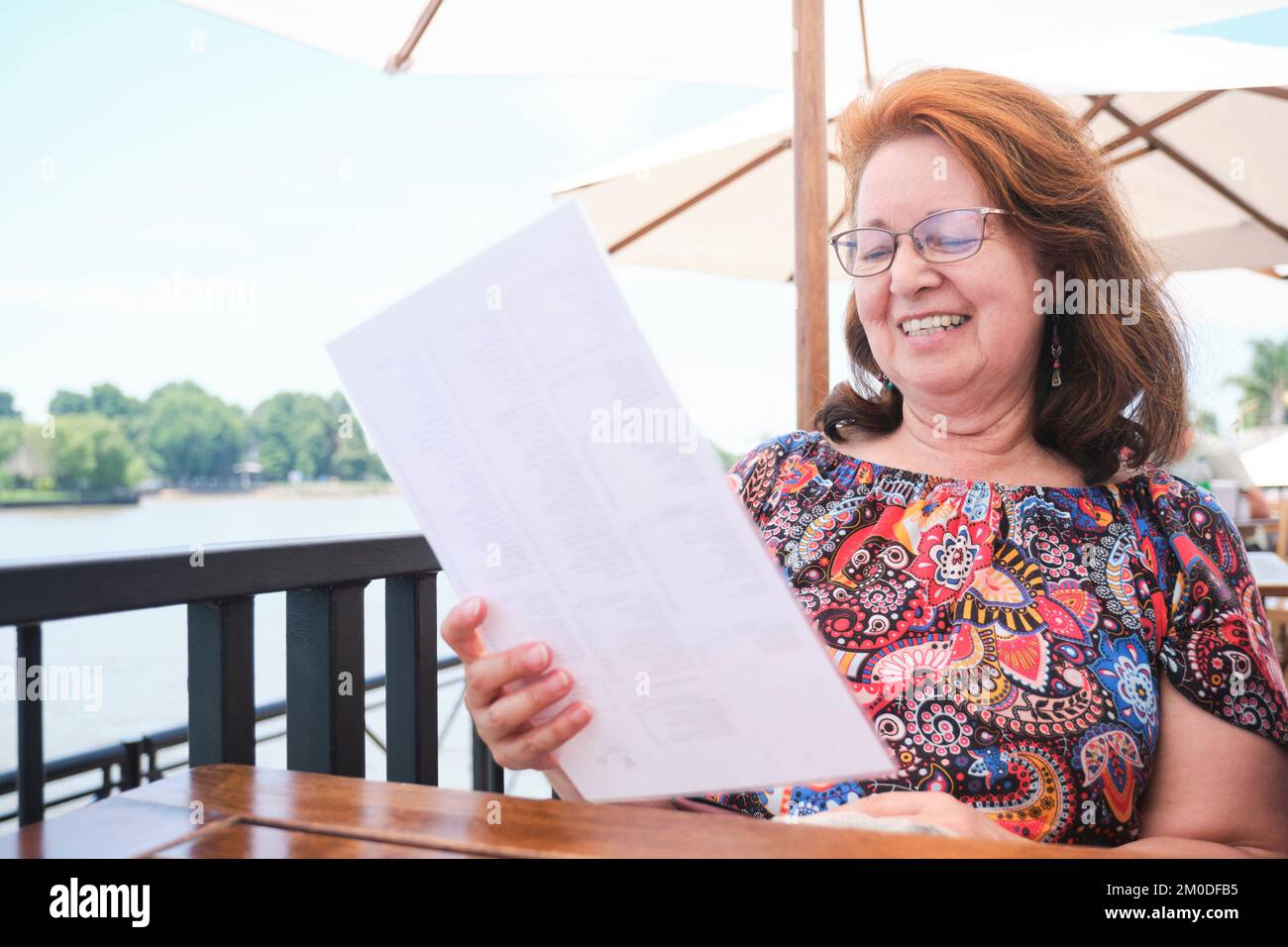 Mature latin woman smiling while reading the menu at an outside table in a restaurant during summer. Stock Photo
