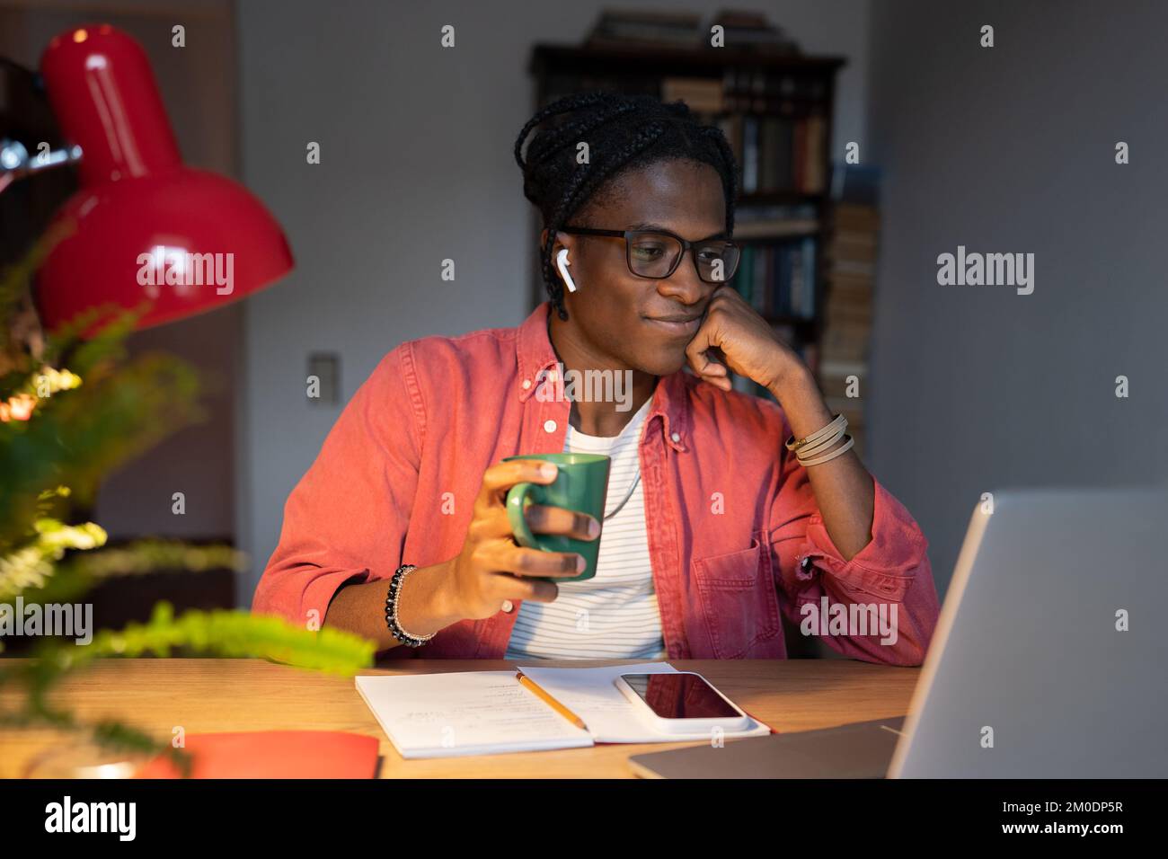 Smiling African student guy in wireless earbuds drinking tea while watching online lectures at home Stock Photo