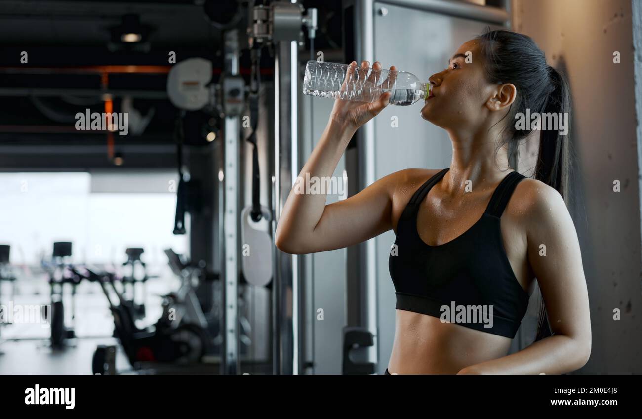 Young woman drinking water from bottle, taking break from exercise at gym. Stock Photo