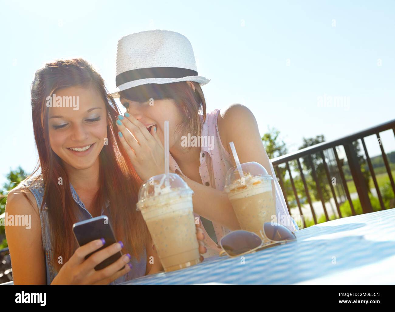Have you heard...A teenage girl texting on a cellphone while her friend whispers something in her ear. Stock Photo