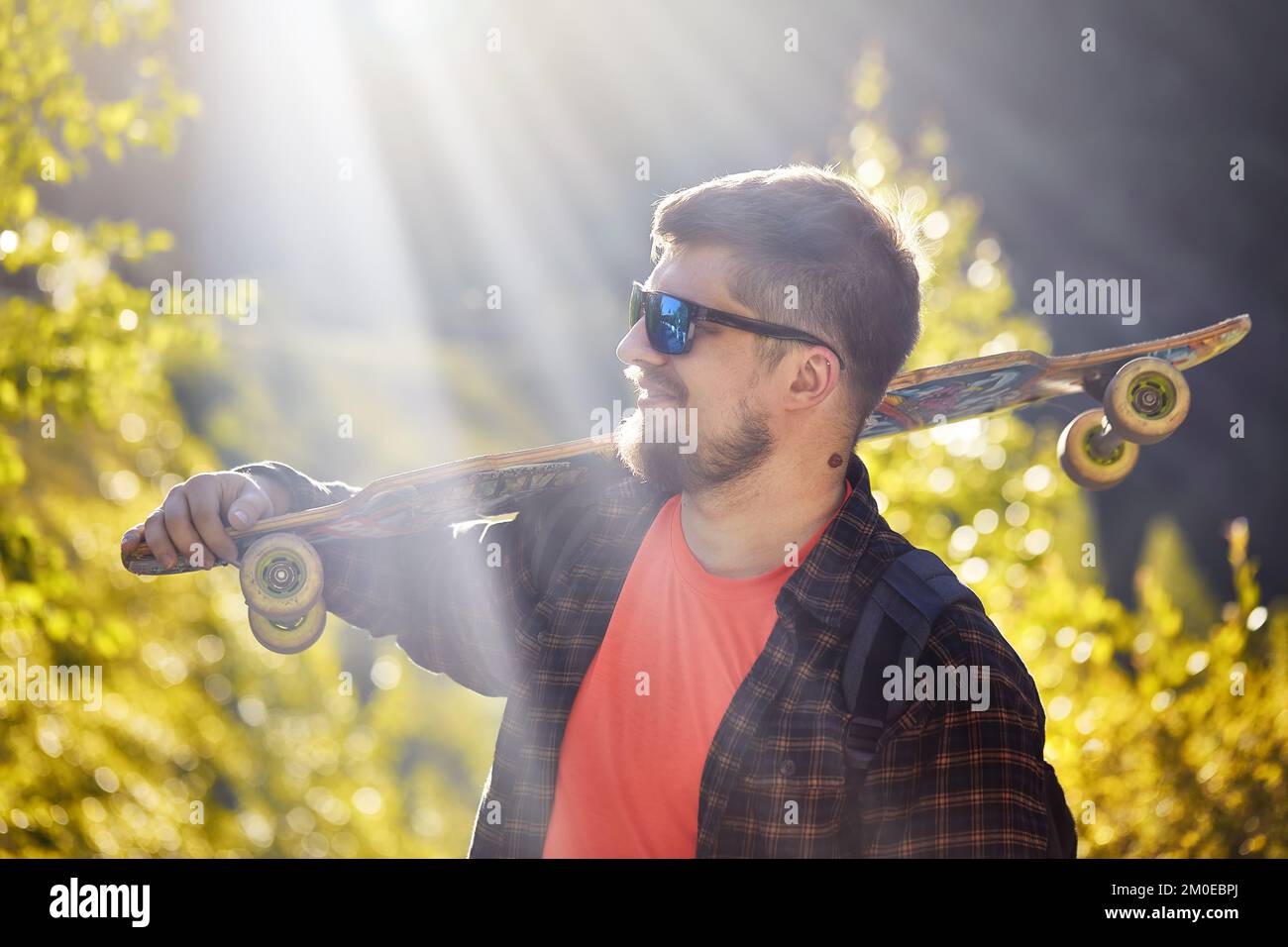 Portrait of serious bearded man Skater with sunglasses holding his longboard in the mountain forest and glowing sunrise rays background Stock Photo