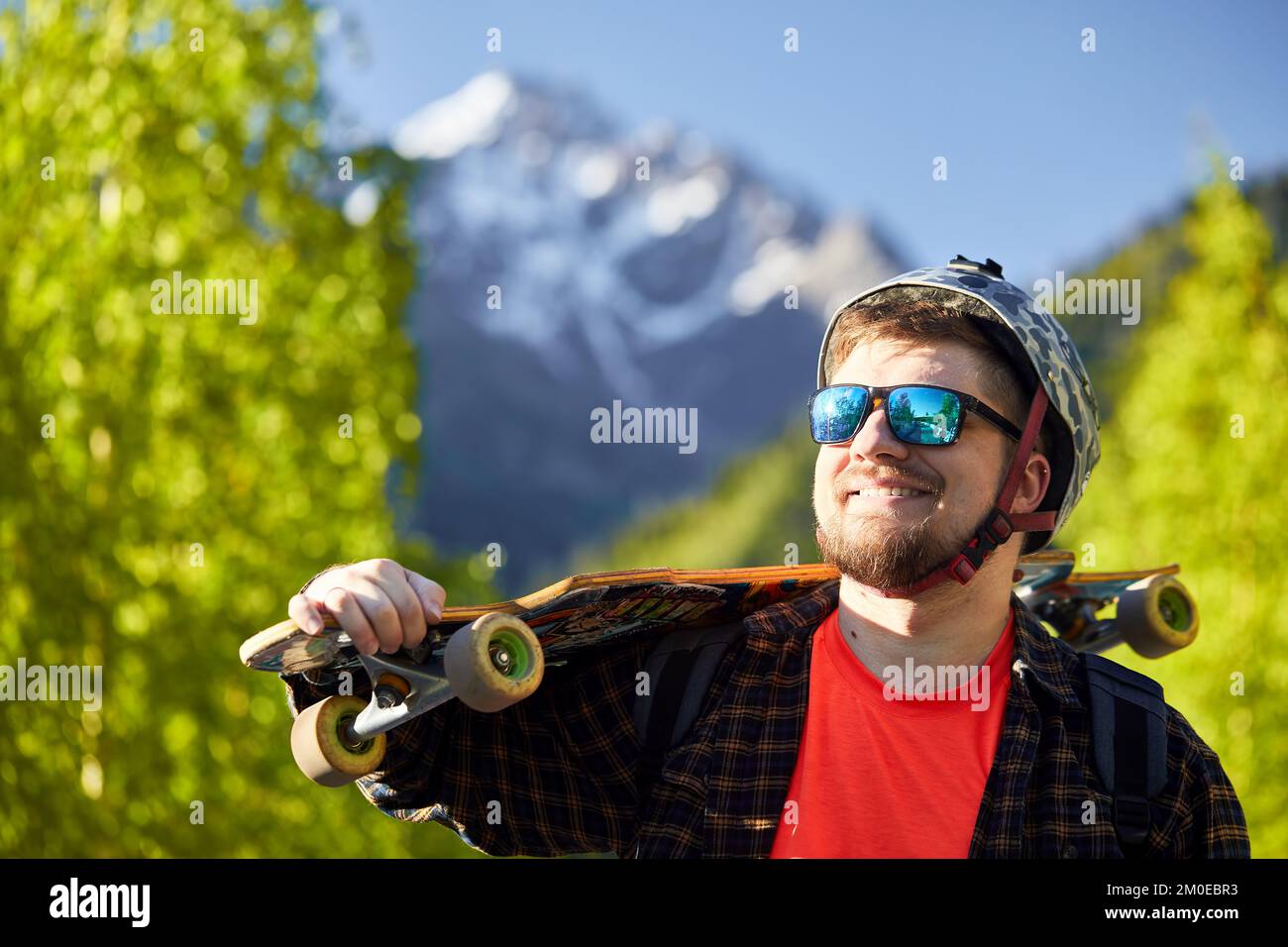 Portrait of happy bearded man Skater with sunglasses and helmet holding his longboard in the mountain at background Stock Photo