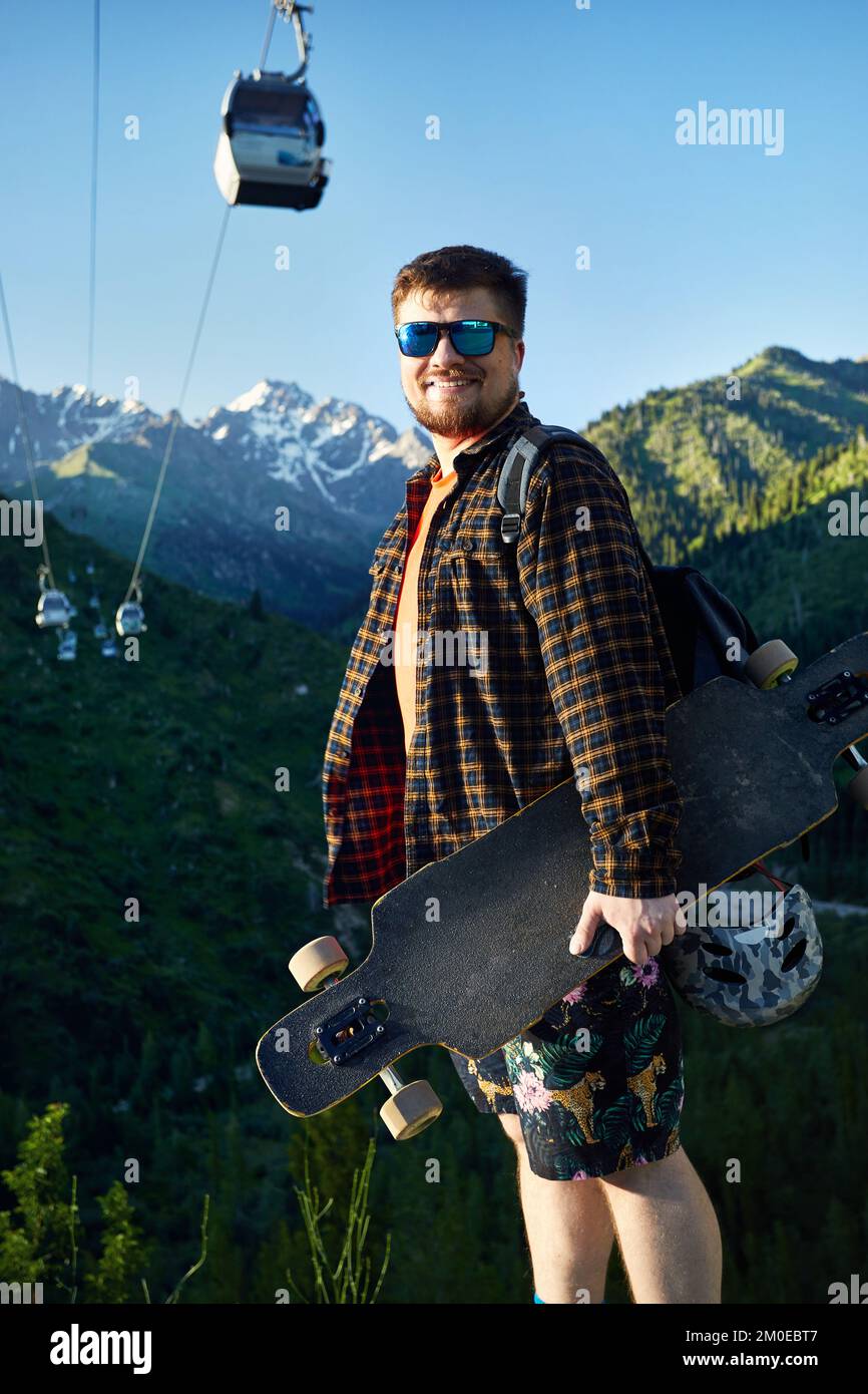Portrait of Bearded man Skater with sunglasses holding his longboard in the mountain road with cable car at background Stock Photo
