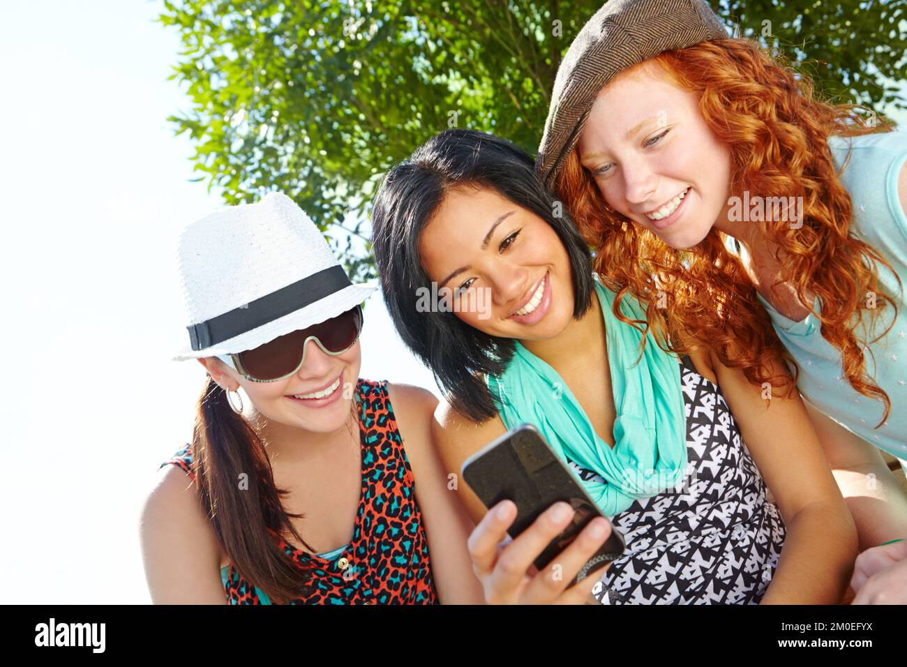 Meet us at the park...Three teenage girls sitting outside with a cellphone smiling at a text message. Stock Photo