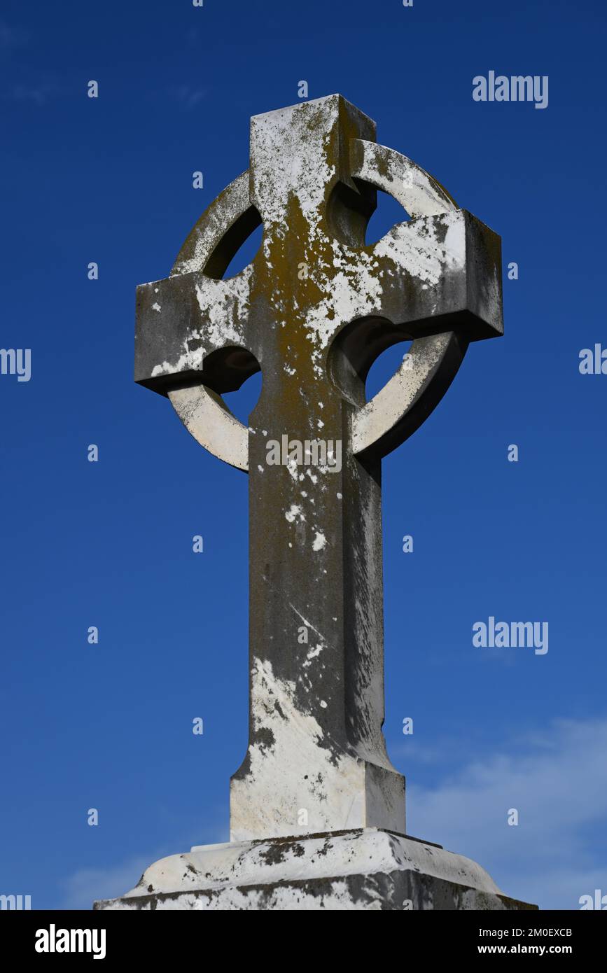 Worn and weathered stone Celtic Cross, originally white but featuring large sections where grey is revealed, with blue sky in the background Stock Photo