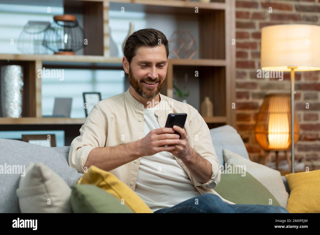 A mature man with a beard in a shirt at home is sitting on the sofa in the living room, using the phone, typing messages and browsing the Internet online. Stock Photo