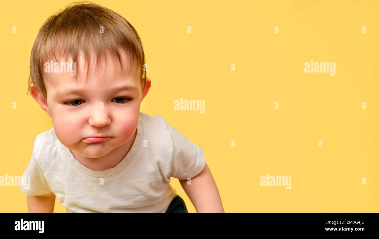 Portrait of a doubting toddler baby on a studio yellow background. Skeptical child sitting on the floor in a white t-shirt and blue jeans, copy space. Stock Photo