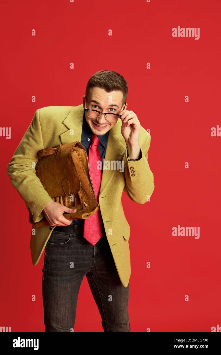 Portrait of young man in stylish jacket, glasses and vintage briefcase posing over red background. New worker, trainee. Shy look. Concept of business Stock Photo