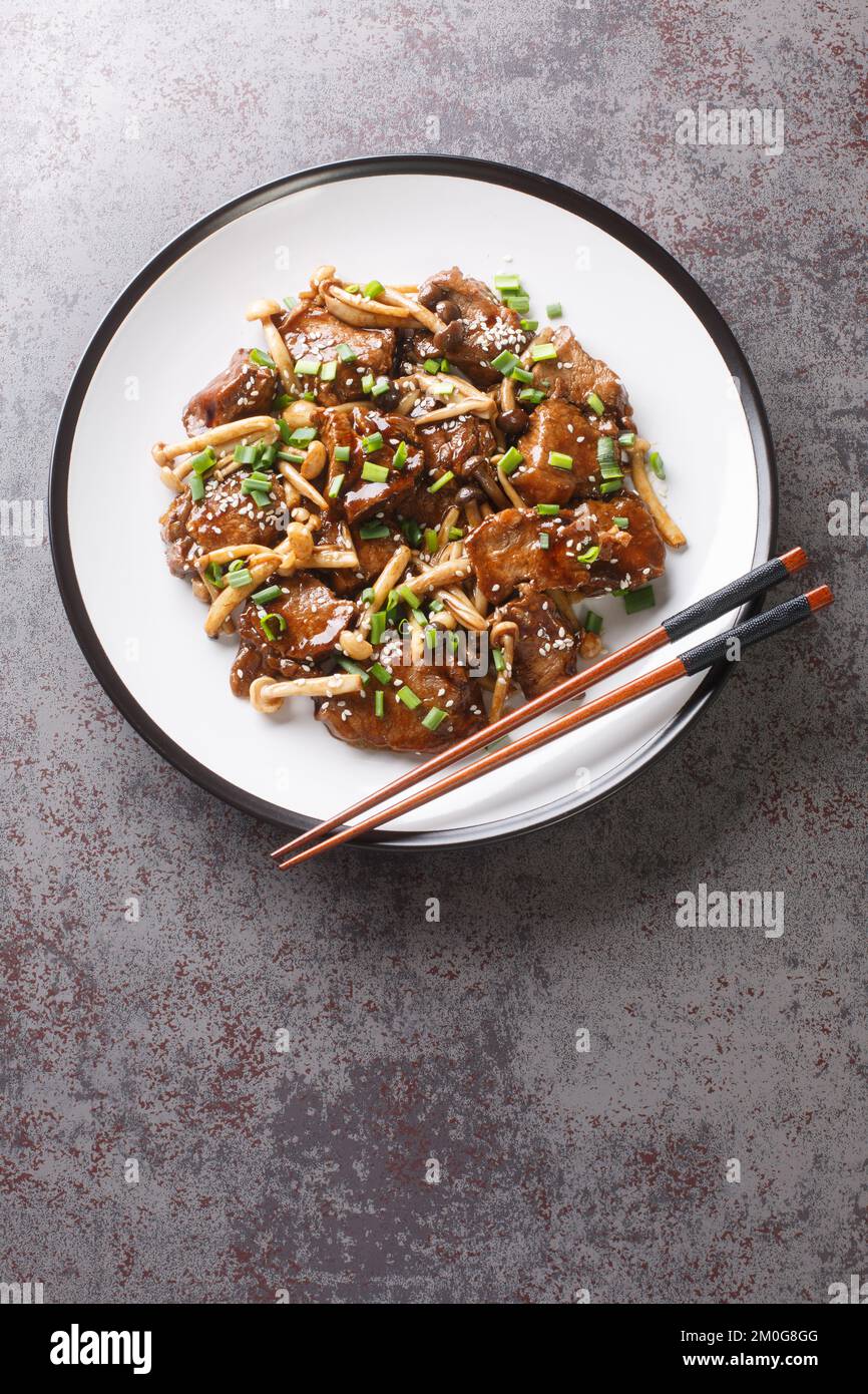 Beef steak with beech mushrooms in teriyaki sauce sprinkled with green onions and sesame seeds close-up in a plate on the table. Vertical top view fro Stock Photo