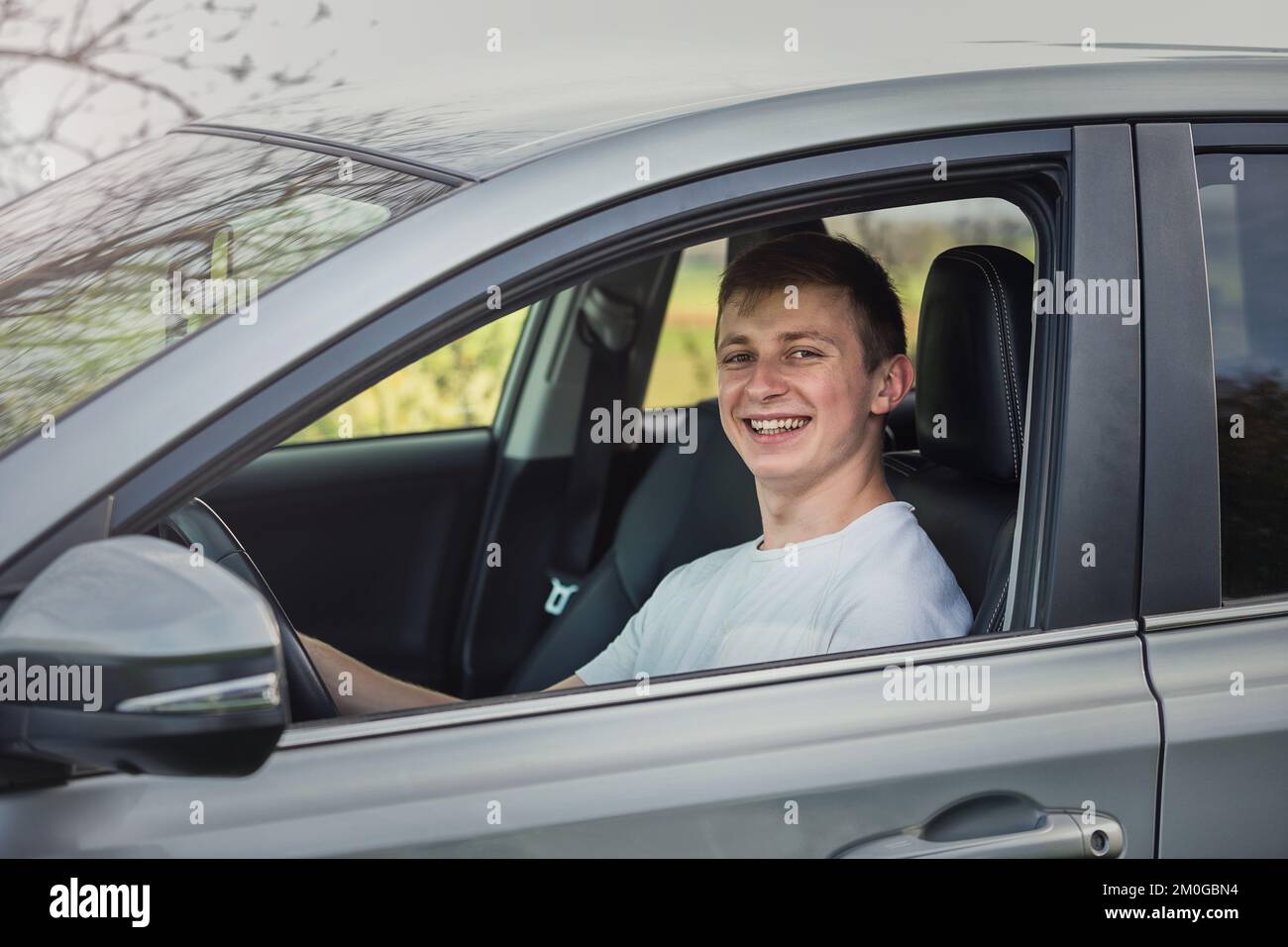 Joyful young man driving safe his new car, looking cheerful to camera