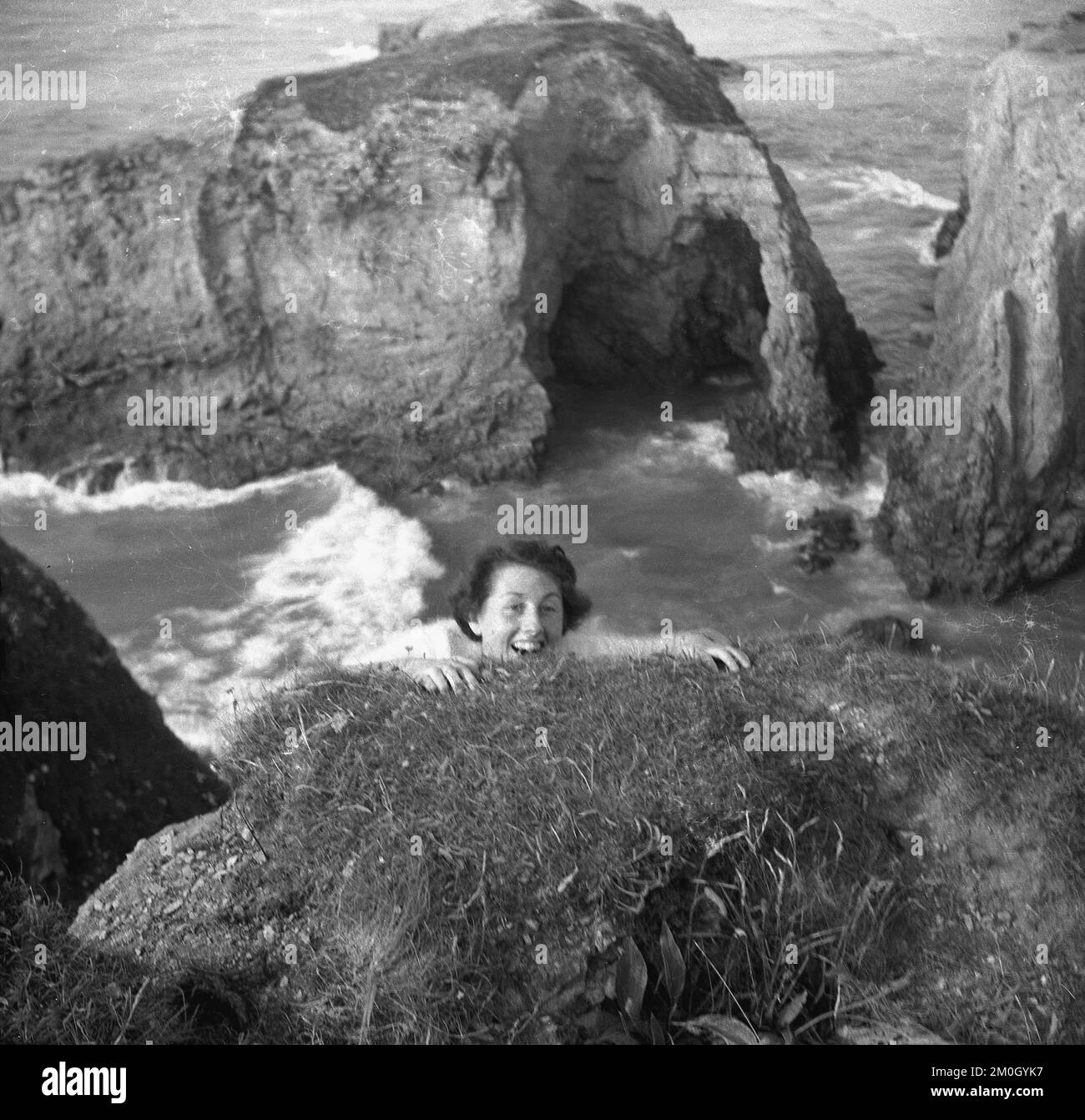 1950s, historical, at the coast, high above the ocean, looking down at the sea and protruding rocks, a lady below the ground, pretending perhaps that she is clinging on to the cliff face or overhang, England, UK. Stock Photo