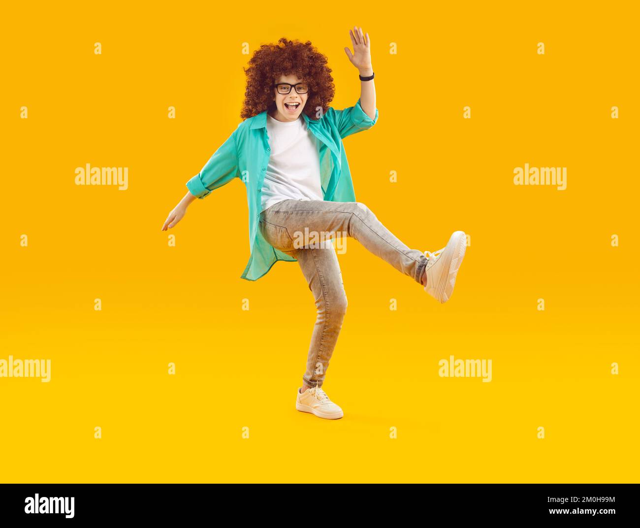 Happy cheerful funny boy in red curly wig, dressed in casual clothes, fooling around and dancing in cheerful mood. Stock Photo