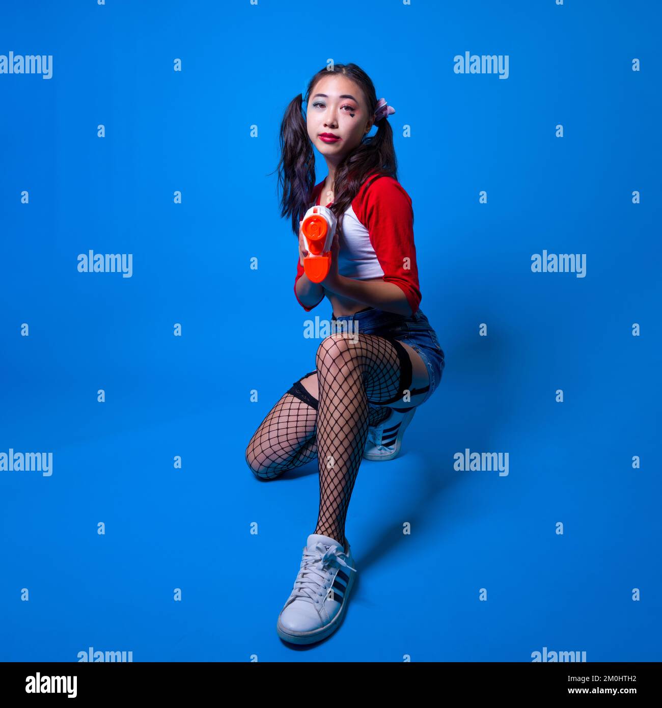 Young Woman in Harlequin Costume Squatting with Water Pistol in Hands on Solid Blue Background Stock Photo