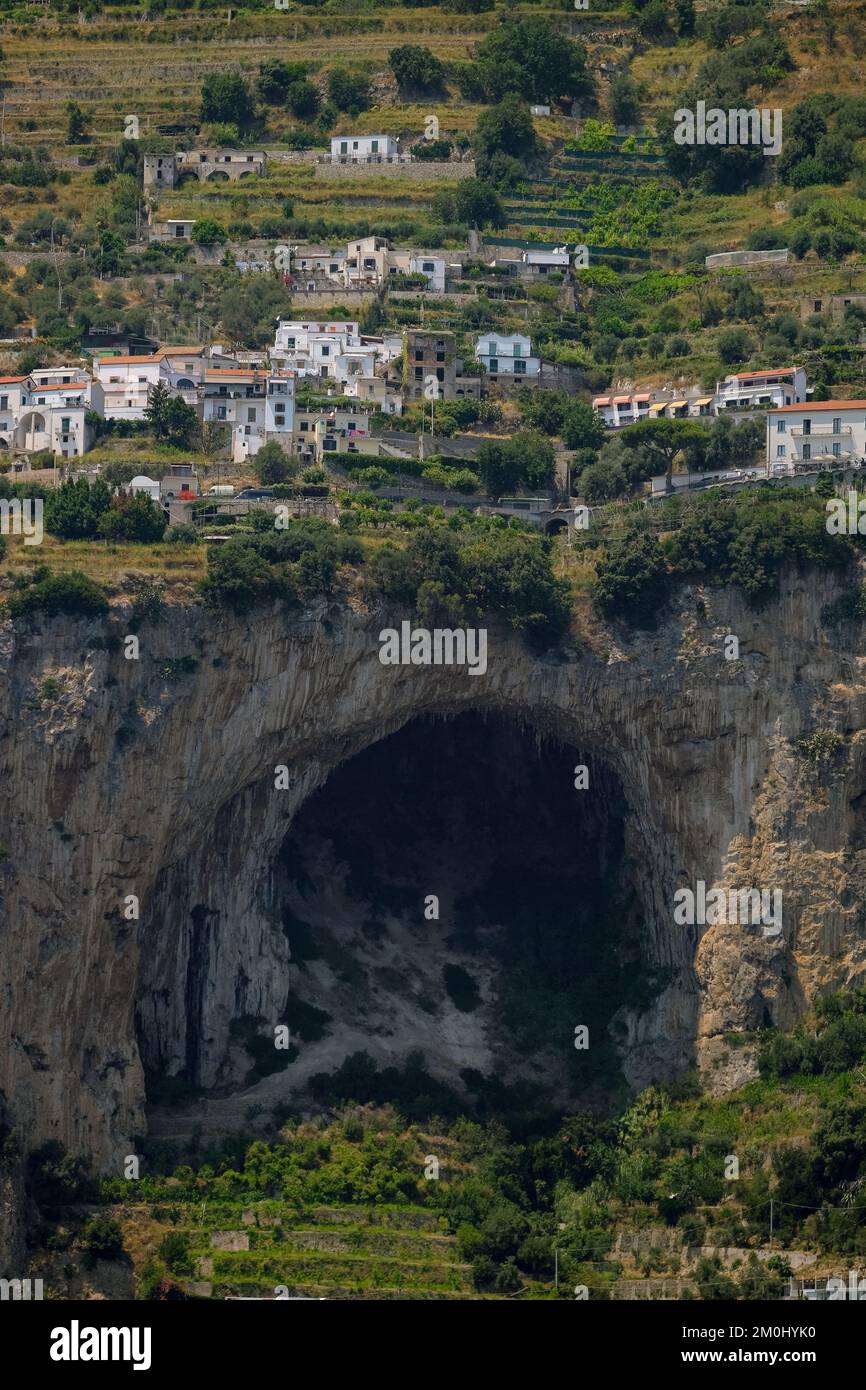 Large cave opening sits on the Amalfi coast amongst the limestone cliffs with houses perched scarily on top with hotel and beach front below. Stock Photo