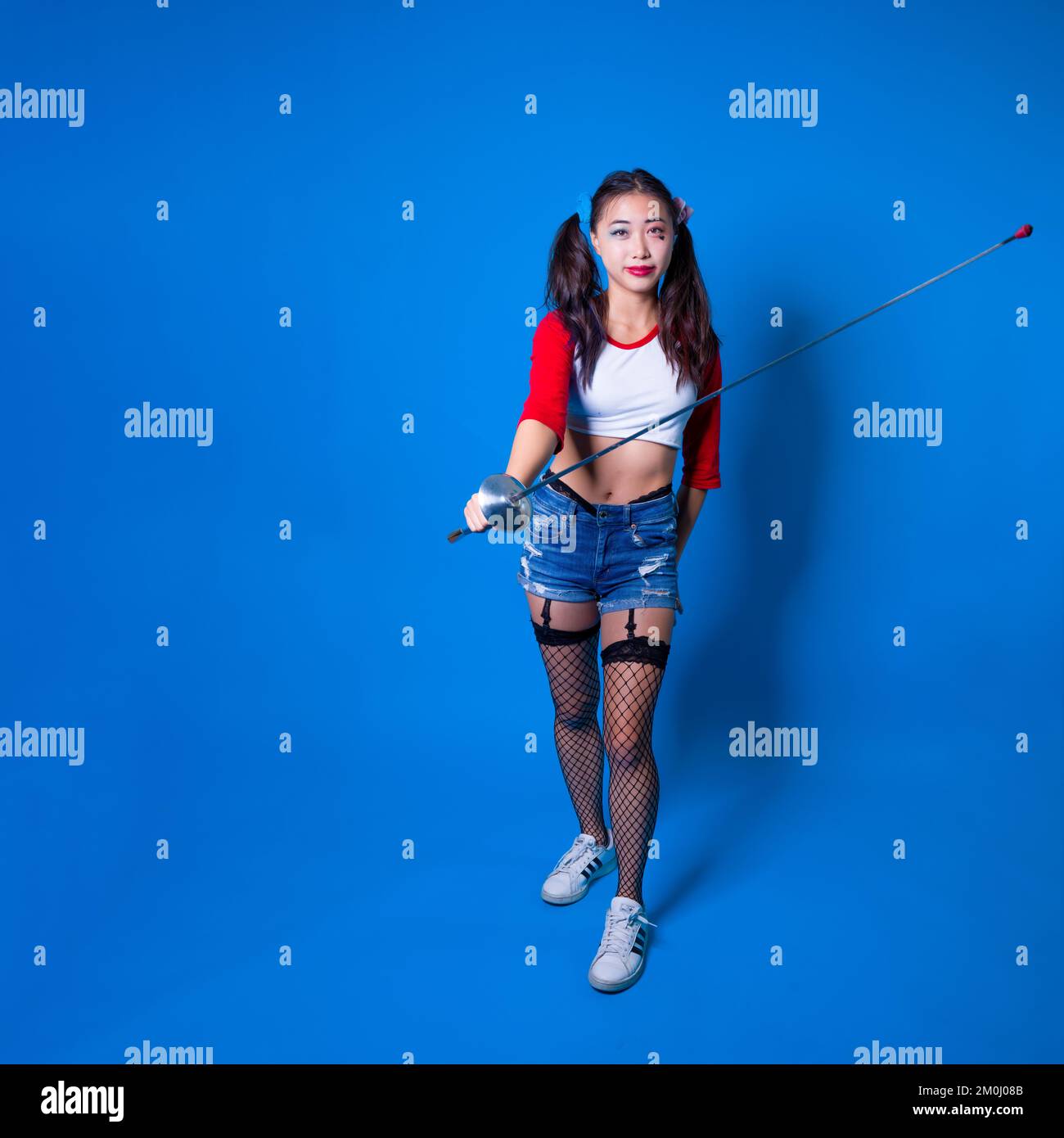 Young Woman in Harlequin Costume Holding Fencing Foil Against a Solid Blue Backdrop Stock Photo