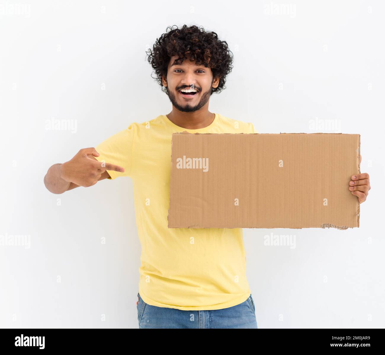 Young Asian man with curly hair and casual stylish clothes smiling, holding a piece of cardboard box and finger pointing Stock Photo