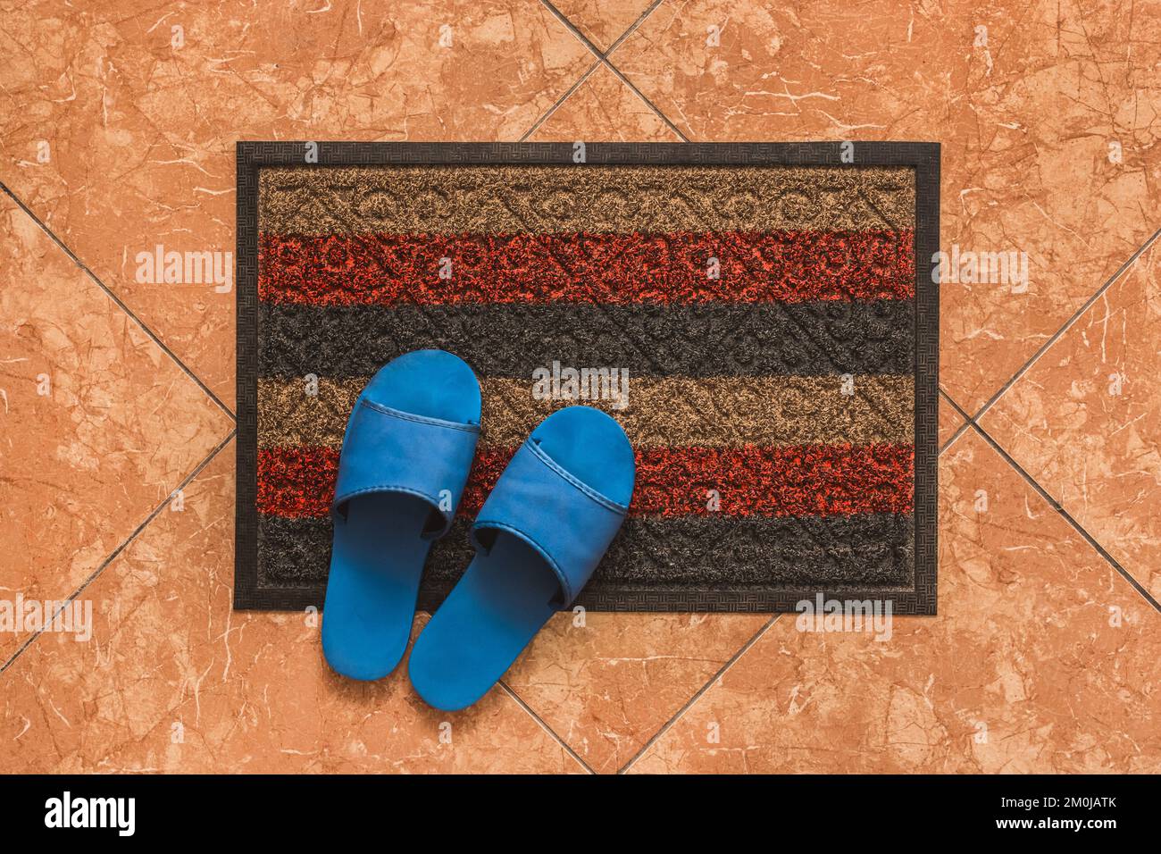 Blue homemade slippers stand on the foot carpet against the background of the floor tiles, the view from above. Stock Photo
