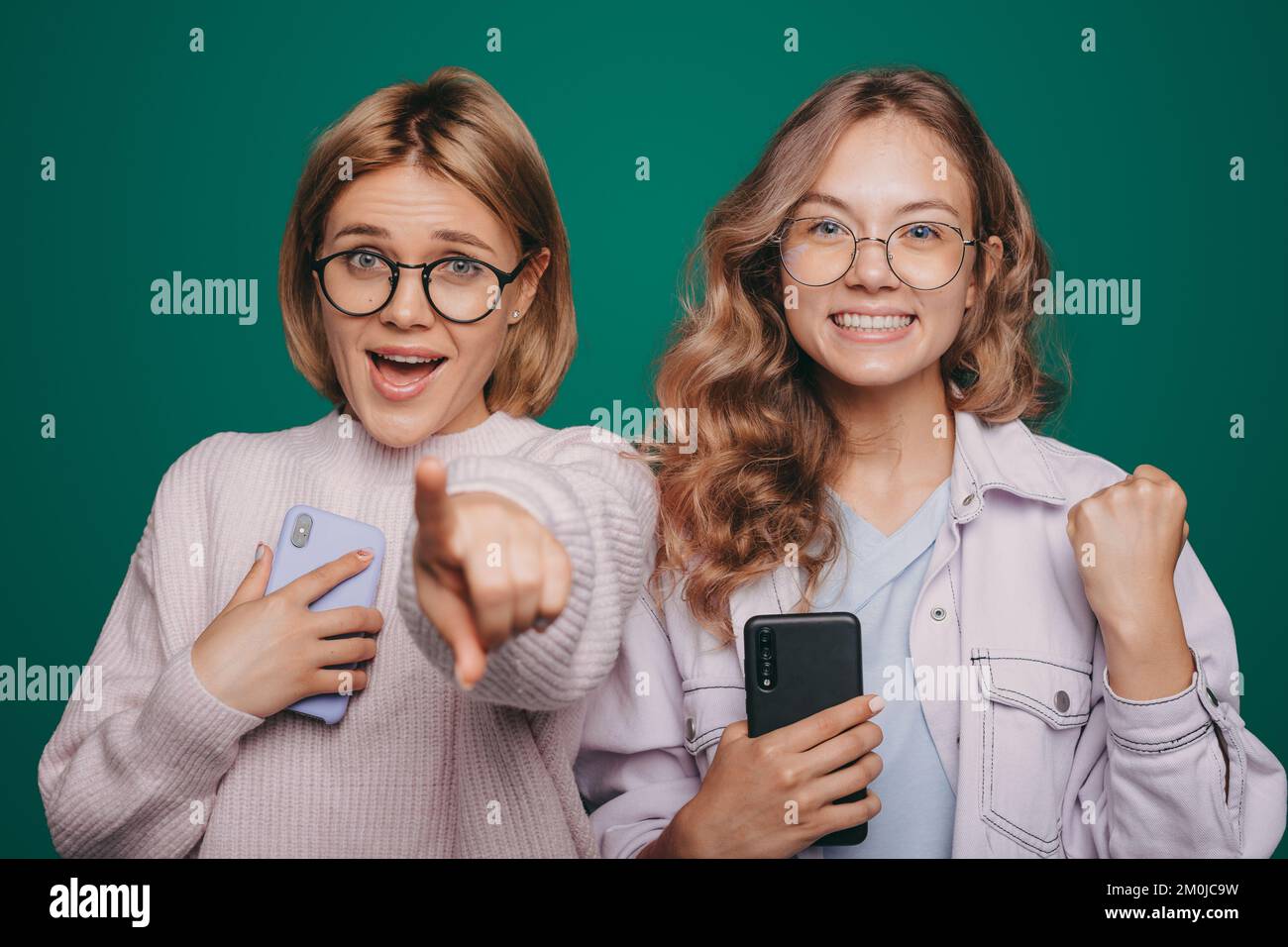 Two satisfied young women friends using mobile cell phone isolated on green background studio portrait. Isolated portrait. Excited cheerful. Stock Photo
