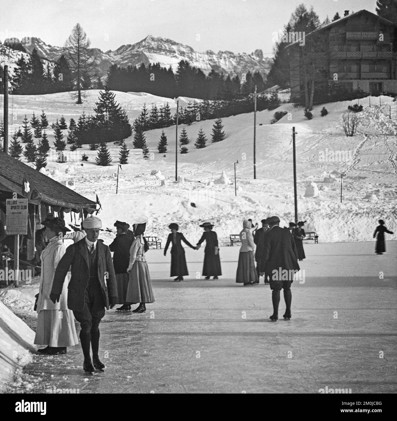 An early 20th Century vintage black and white photograph taken in Sass-Fee in Switzerland, showing a number of people skating on an outdoor skating rink. A sign for The Villars Sports Club on the left. Stock Photo
