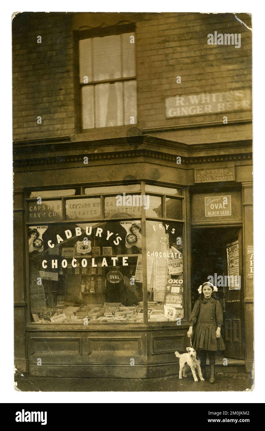 Original pre WW1 postcard of young girl called Gladys, with a pet dog terrier, standing outside her parent's tobacconists shop front  - W. Young tobacconist. The shop also sells sweets and there's a Cadbury's chocolate sign in the window. The shopkeeper peers through the window. Posters for the silent films in the shop window for 'Out of the Deep' 1912 silent film, 'Buckskin Jack' 1911, Four Singing Miners (named this up to 1912) Photo dated 1912. There's a West Bromwich football fixtures advert which indicates the town was West Bromwich, Staffordshire, West Midlands, England, UK. Stock Photo