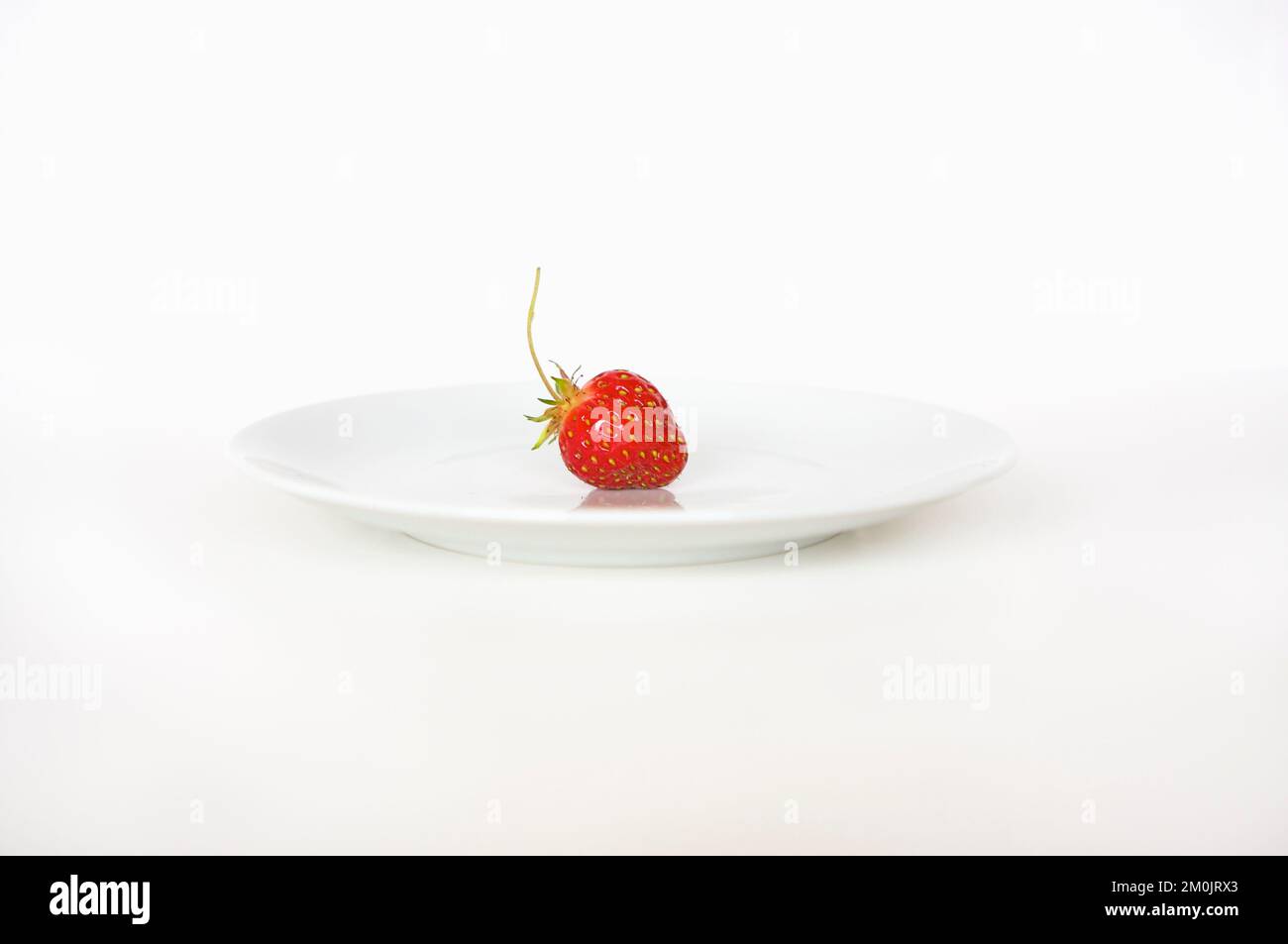 A single ripe red strawberry sitting on a white plate against a bright, clean white background with lots of copy space. Stock Photo